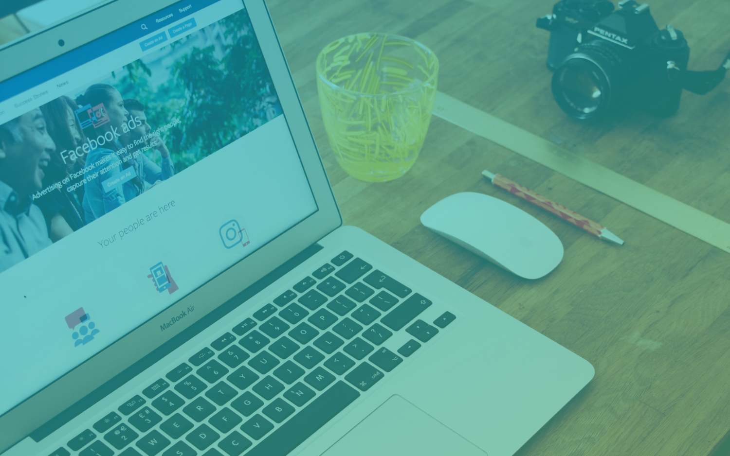 Image of a laptop sitting on a desk next to a yellow cup of water, a mouse, pencil, and camera